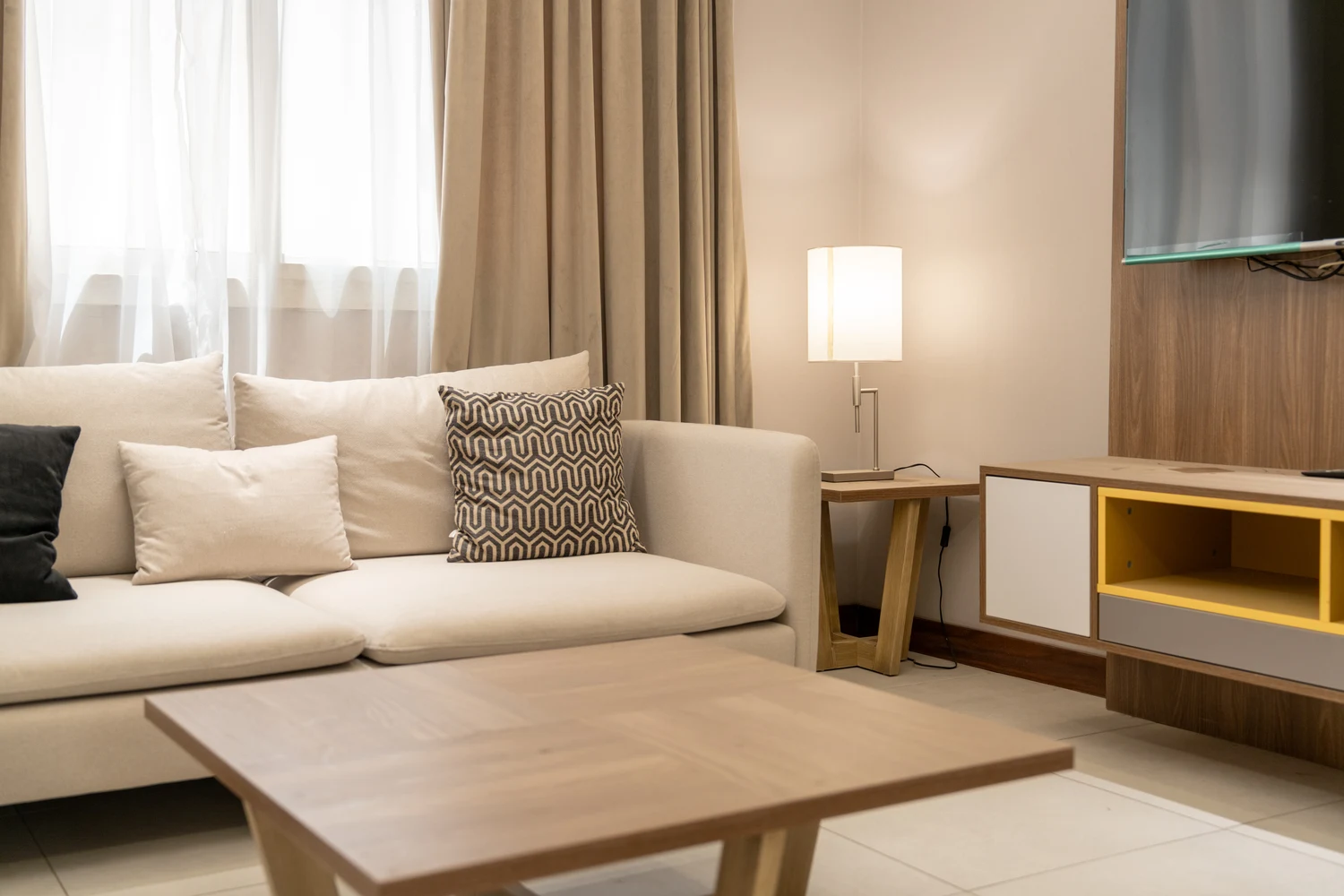 Abraj INN Suites, Doha Hotels: cream corner sofa, a curtained window, a lit table lamp and a wall-mounted TV.